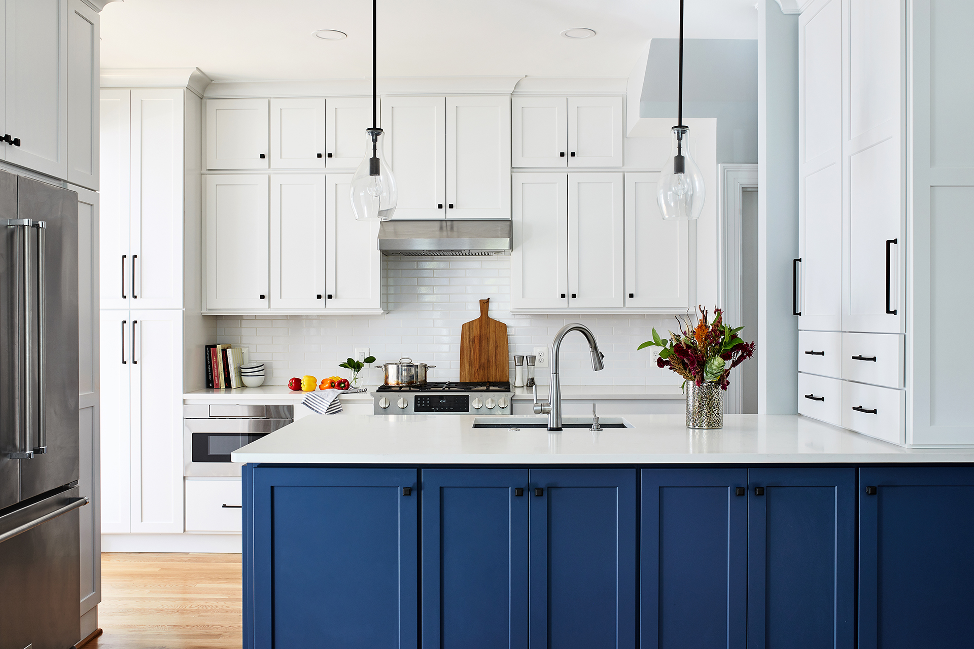 white and blue kitchen with stainless steel appliances, two bell pendant lights, kitchen island with farmhouse sink and floating white cabinets with black knobs
